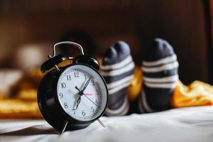 reloj que muestra las 7 en punto en la cama con los pies de una persona dormida en calcetines calientes en el fondo foto