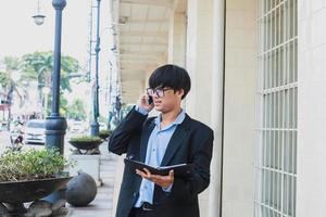 Young asian wearing black suit and glasses doing call by phone while holding a note book and looking on the street photo