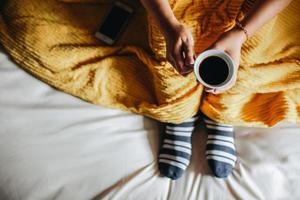A people wearing socks under blankets on the bed and holding a cup of coffee