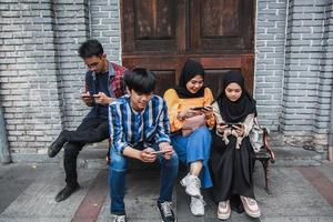 Four young friends sitting outdoors and looking at mobile phone. Group of people sitting on a bench and playing online games on smart phone photo