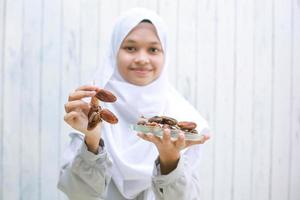 Young asian muslim female  smiling and offering dates on her hand while holding dates on the plate photo