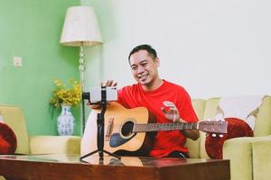 Smile asian man holding a guitar with say hi gesture while looking smart phone during live streaming video photo