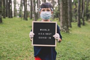 Asian boy wearing face masks and rubber hand gloves standing while holding quote on letter board says World Against Covid 19 photo