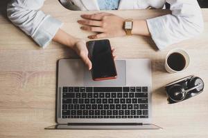 Top view of woman hand are holding and using blank screen smartphone with laptop, coffee and clock on a wooden office desk photo