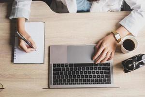 Top view of woman hand are writing on the book and using a laptop computer with coffee and clock on a wooden office desk photo