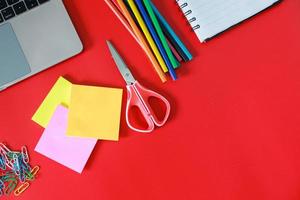 Top view of office or school supplies isolated on red background. Open book, colored pencil, laptop, scissor, colorful paper clip and sticky note with free or copy space photo