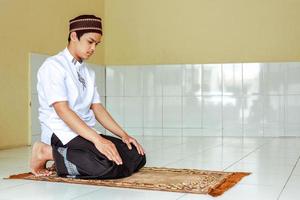 Religious muslim man praying in the mosque while wearing islamic clothes with pose of sitting between two prostrations photo