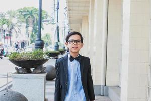 niño asiático feliz con traje negro vintage y gafas posando y mirando a la cámara foto