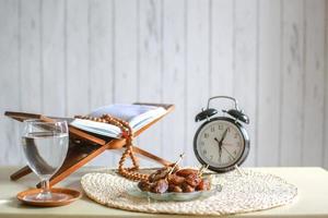 Iftar time cincpet. Kurma or dates fruit with glass of  water, holy Quran, alarm clock showing 6 o'clock and prayer beads on the table. photo