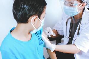 Doctor wearing complete PPE with syringe needle inject to boy patient. Medicine, vaccination, immunization and health care concept photo
