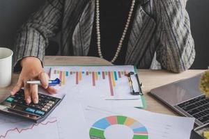 Cropped shot of business woman using  calculator with graph and chart on messy office desk. Financial report, business accounting, graph finance, analysis data charts photo