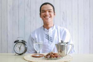 hombre musulmán vestido con koko sonriendo a la cámara con fechas, reloj, tetera y agua llena de vidrio sobre la mesa. Ramadán tradicional, hora de iftar. concepto de mes de ayuno de ramadán kareem con enfoque selectivo foto