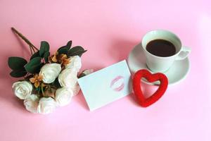 Cup of coffee with rose, kiss marked on white envelope and heart shape isolated on pink background photo