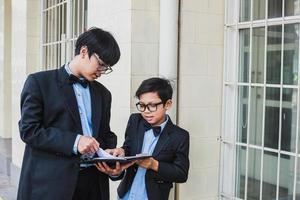 Two teenager wearing glasses, black ribbon and vintage black suit jacket standing while holding and reading a note book photo