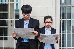 Two teenager wearing glasses, blue shirt and black suit jacket reading newspaper photo