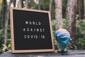 Earth day concept Quote on letter board says World Against Covid 19 and Earth globe in sterile medical mask with blurred nature background. photo