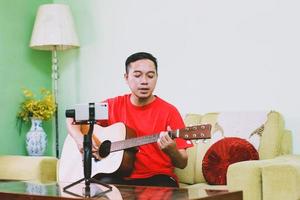 Portrait of asian man singing and playing a guitar while recording by smart phone at home photo