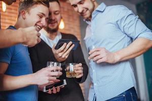 Cheerful old friends communicate with each other and phone watch,  glasses of whiskey in pub. Concept  entertainment  lifestyle. Wifi connected people in bar table meeting photo