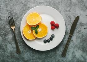 postre saludable plato blanco con rodajas de naranja, frambuesas, arándanos y cubiertos antiguos