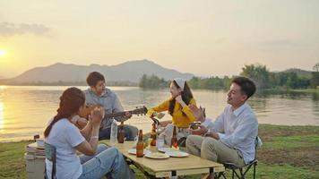 Group Four People Friends Asian men and women Camping, drinking beer, celebrating, Singing, Playing Guitar, Having fun and Enjoying Ground tent. Reservoir area during Sunset Vacation Time.
