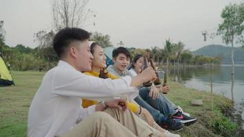 gruppo asiatico quattro giovani amici uomini e donne che fanno un picnic in campeggio vicino al fiume, chiacchierano, ridono, bevono festeggiano birra allegria. video