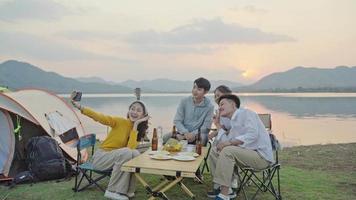 gruppo quattro persone amici uomini e donne asiatiche picnic in campeggio festeggiando, selfie tramite smartphone divertendosi e godendosi la tenda a terra. area del serbatoio durante le vacanze al tramonto. video