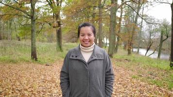Front view of Asian woman standing at the forest, smiling and looking at camera. Autumn season, leaves falling from the trees , beautiful nature, getting some fresh air at the forest, Sweden video