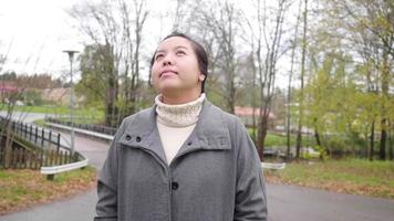 Front view of Asian woman standing and looking around the park, Autumn season, leaves falling from the trees , beautiful nature, getting some fresh air at the park, Sweden video