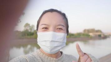 närbild av glad asiatisk kvinna som står och tar videoselfie vid floden. bär mask. flod och träd bakgrund, ser vacker utsikt video