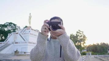 vista frontal da mulher asiática em pé e tirando foto no parque em lindo dia. segurando a câmera. árvores e fundo de estátua de monge. dia relaxante à noite video