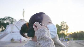 närbild av asiatisk kvinna som står och andas i parken, tar ut masken och får lite frisk luft. stort träd och munk staty bakgrund video