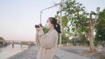 femme asiatique debout et prenant une photo de la vue sur la rivière le soir tenant un appareil photo avec fond de parc en journée de détente video