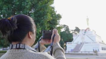 vista trasera de una mujer asiática parada y usando una cámara para tomar una foto en el parque en un hermoso día. gran árbol y fondo de estatua de monje. día relajante, felices vacaciones. video