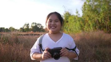 Front view of happy Aisan girl standing in meadow and holding a camera, smiling and looking at camera. Standing on grass in beautiful day with blue sky background video