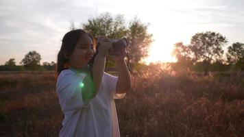 Fröhliches asiatisches Mädchen, das mit der Kamera auf der Wiese ist und abends ein Bild von der Aussicht auf einen Bauernhof macht. Stehen auf Gras an einem schönen Tag mit blauem Himmelshintergrund. wunderbares Sonnenlicht video