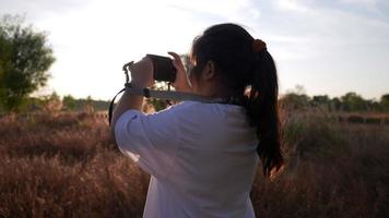 vista posteriore della ragazza asiatica felice che è nel prato con la macchina fotografica, scattando una foto di vista la sera. in piedi sull'erba in una bella giornata con sfondo azzurro del cielo. meravigliosa luce del sole video