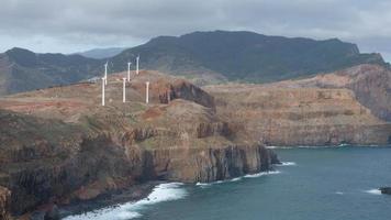 Windmills on the hills next to the coast. Renewable energy, green energy. Ocean and waves. Madeira Island in Portugal. Cloudy day and cliffs with brown color. video