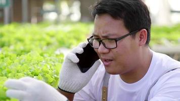 primo piano contadino occhiali uomo serio parlando con il cliente tramite smartphone in serra idroponica. problema di negoziazione dello stress aziendale agricolo in un'azienda agricola biologica video