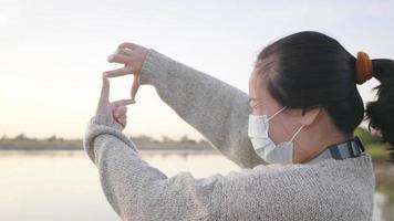 cerrar la vista trasera de una mujer asiática de pie y mirando la vista a través del río, usando una máscara. fondo de río y árboles, con una hermosa vista video