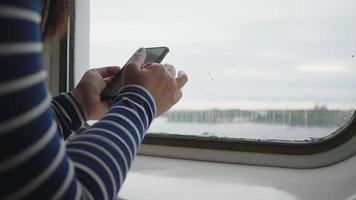 nahaufnahme hand mit smartphone am fenster, schiebebildschirm auf dem smartphone. Sitzen auf dem Boot am Fenster mit schöner Aussicht nach draußen video