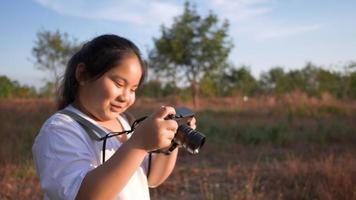 menina asiática feliz no Prado com a câmera, tirando uma foto de vista. de pé na grama em lindo dia com fundo de céu azul video