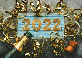 fondo del concepto de celebración de la víspera de año nuevo. máscara médica con los números 2022 y botella de champán con gafas. concepto de año nuevo covid-19 sobre fondo gris foto