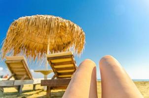 Woman girl legs sunbathing on tropical sandy beach with blurred straw sunshade umbrella and wooden sun lounger photo