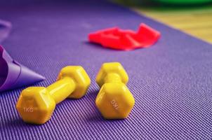 Sports equipment close-up view, yellow dumbbells and red rubber resistance band on violet ribbed yoga mat photo