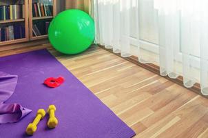 Sports equipment on floor in living room, violet yoga mat, yellow dumbbells, red rubber resistance band and green rubber fitness aerobic ball photo