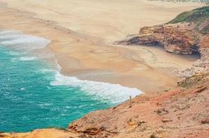 vista aérea superior de la playa de arena con rocas y acantilados y olas de agua turquesa azul del océano atlántico cerca de la ciudad de nazare foto