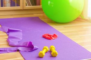 Sports equipment on floor in living room, violet yoga mat, yellow dumbbells, red rubber resistance band and green rubber fitness aerobic ball photo