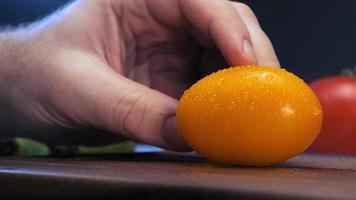 Person hand holds yellow tomato with water drops and sharp knife cuts vegetable into slices under electric light extreme closeup video