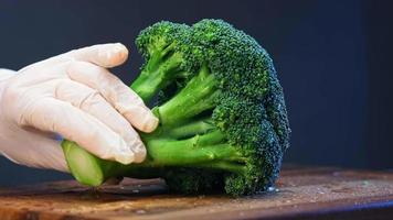 Vegan holds broccoli and cuts into small pieces with knife on brown wooden board under bright electric light extreme closeup video