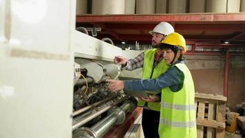 i lavoratori in uniforme di sicurezza e gli ingegneri industriali con elmetto protettivo utilizzano il computer portatile per controllare e controllare le macchine. due professionisti lavorano nella fabbrica di produzione della carta, mantengono le apparecchiature di produzione. video